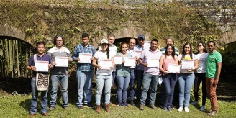 Course participants stand together on grass for a photo in front of a moss-covered bridge, proudly presenting their course completion certificates and smiling.